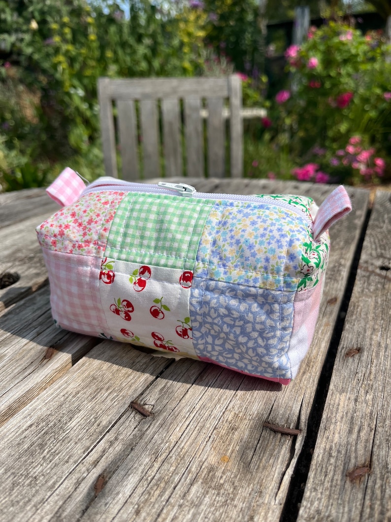 K&ouml;nnte beinhalten: Eine kleine Patchwork-Kosmetiktasche mit einem wei&szlig;en Rei&szlig;verschluss. Die Tasche besteht aus verschiedenen Stoffen mit Blumen-, Karomuster- und Kirschmotiven in Rosa, Gr&uuml;n und Blau.