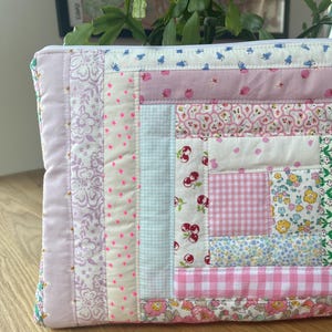 May include: A pink and white patchwork zippered pouch with a variety of floral and geometric patterns. The pouch is made of quilted fabric and has a white zipper.