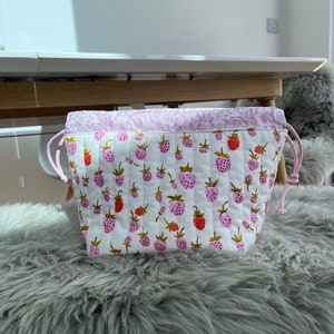 Puede incluir: Una bolsa blanca con cordón y un estampado acolchado de fresas. La bolsa presenta fresas rosas y rojas con hojas verdes. El borde superior tiene un estampado floral rosa y cordones rosas. La bolsa está sobre una superficie gris y esponjosa.