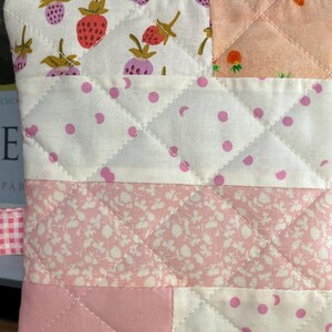 Pu&ograve; includere: Un astuccio trapuntato con cerniera gialla e nastro a quadretti rosa. L'astuccio presenta un design patchwork con vari tessuti, tra cui stampe di fragole e floreali, e tessuto bianco a pois rosa. Le cuciture creano un motivo a rombi.