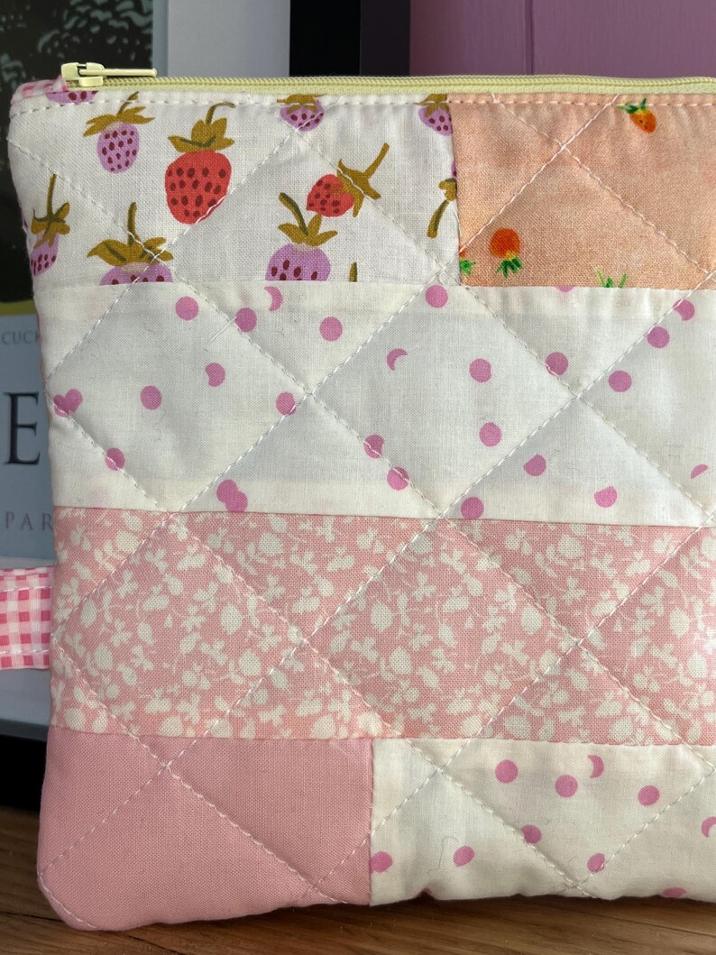 Pu&ograve; includere: Un astuccio trapuntato con cerniera gialla e nastro a quadretti rosa. L'astuccio presenta un design patchwork con vari tessuti, tra cui stampe di fragole e floreali, e tessuto bianco a pois rosa. Le cuciture creano un motivo a rombi.