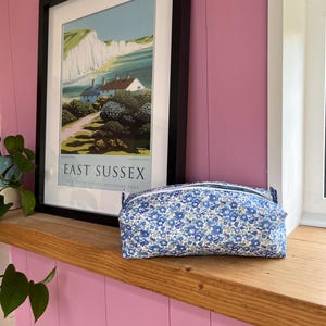 May include: A blue floral print cosmetic bag with a zipper closure sits on a wooden shelf. The bag is in front of a framed poster of East Sussex. The background is a pink wall.