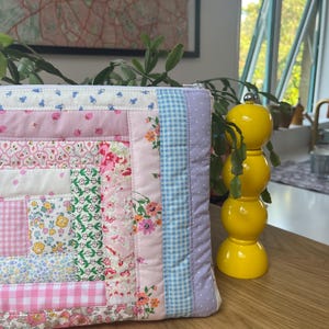 May include: A patchwork quilt-style zippered pouch with a variety of floral and gingham patterns in pink, blue, and white. The pouch is lying on a wooden surface next to a yellow ceramic pepper shaker.