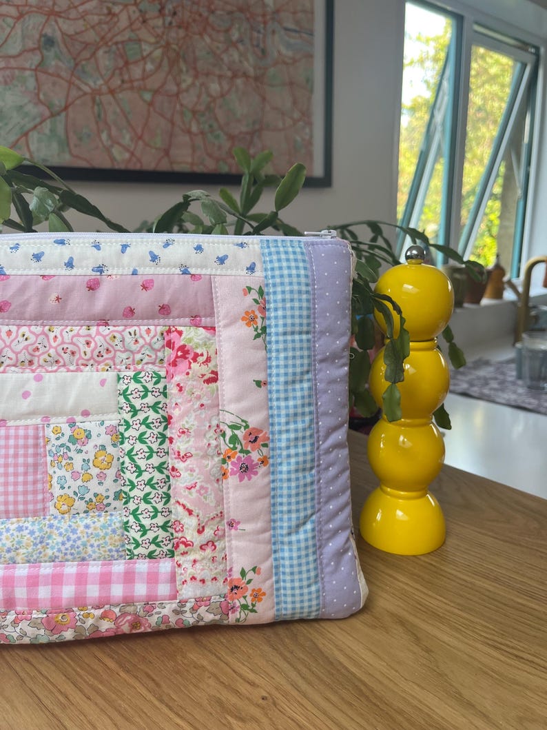 May include: A patchwork quilt-style zippered pouch with a variety of floral and gingham patterns in pink, blue, and white. The pouch is lying on a wooden surface next to a yellow ceramic pepper shaker.