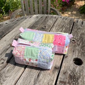 K&ouml;nnte beinhalten: Zwei gepolsterte Beutel mit Rei&szlig;verschluss aus Patchwork-Stoff mit verschiedenen floralen und karierten Mustern in Rosa, Gr&uuml;n, Gelb und Blau.