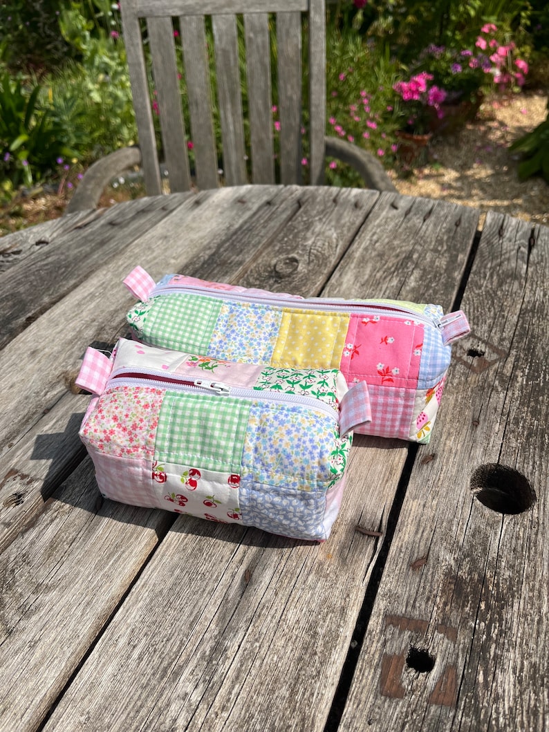 K&ouml;nnte beinhalten: Zwei gepolsterte Beutel mit Rei&szlig;verschluss aus Patchwork-Stoff mit verschiedenen floralen und karierten Mustern in Rosa, Gr&uuml;n, Gelb und Blau.