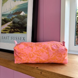 May include: A quilted cosmetic bag in pink and orange. The bag has a floral pattern and diamond stitching. It features a zip closure and small fabric loops. The bag is on a wooden surface. The background includes a framed poster.