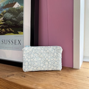 Könnte beinhalten: Kleine, rechteckige Tasche mit hellblauem Hintergrund und weißem Blumenmuster. Die Tasche hat einen Reißverschluss und liegt auf einer Holzoberfläche. Der Hintergrund umfasst ein gerahmtes Poster und eine rosa Wand.