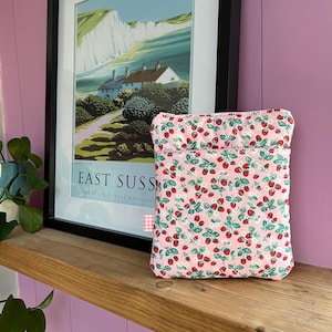 May include: A pink quilted pouch adorned with a strawberry print, displaying red strawberries, green leaves, and white flowers. The pouch rests on a wooden shelf, with a framed landscape print in the background. The print includes the text "EAST SUSSEX".
