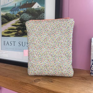 May include: A rectangular floral print pouch with a pink zipper and a small pink and white checkered tag. The pouch is filled with a colorful floral pattern on a white background. The pouch is sitting on a wooden shelf.