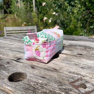 K&ouml;nnte beinhalten: Eine kleine rosa und gr&uuml;ne Patchwork-Stofftasche mit Rei&szlig;verschluss. Die Tasche liegt auf einem Holztisch.