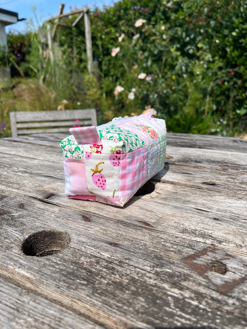 K&ouml;nnte beinhalten: Eine kleine rosa und gr&uuml;ne Patchwork-Stofftasche mit Rei&szlig;verschluss. Die Tasche liegt auf einem Holztisch.