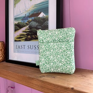 May include: A small, rectangular, green floral print pouch with a zipper closure. The pouch is made of a light green fabric with a white floral pattern. A small green and white checkered tag is attached to the side. The pouch is sitting on a wooden shelf.