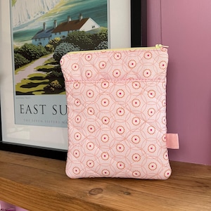 May include: A pink fabric pouch with a repeating pattern of orange circles and pink dots. The pouch has a yellow zipper and a small pink fabric tag. The background includes a framed poster and a pink wall.