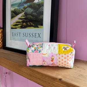 May include: A patchwork fabric pouch with a pink zipper and small handles. The pouch is made of various patterned fabrics, including floral, polka dot, and gingham. The background features a framed poster with the text "EAST SUSSEX".