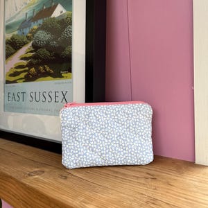 Può includere: Un piccolo astuccio rettangolare trapuntato con un motivo floreale azzurro e bianco. L'astuccio ha una cerniera rosa ed è appoggiato su una superficie di legno. Un poster incorniciato con la scritta "EAST SUSSEX" è sullo sfondo.