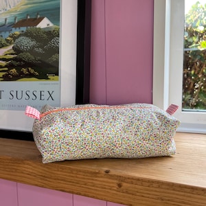 May include: A rectangular floral print pouch with a pink zip and gingham tabs. The pouch is filled and sits on a wooden surface. The floral pattern includes small yellow, pink, and blue flowers on a white background.