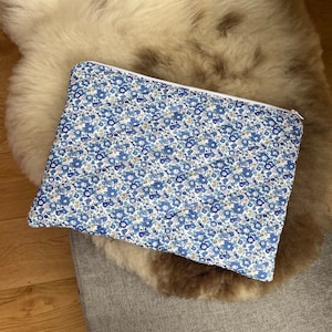 Könnte beinhalten: Eine blaue und weiße, mit Blumen gemusterte Reißverschlusstasche. Die Tasche ist gesteppt und hat einen weißen Reißverschluss.