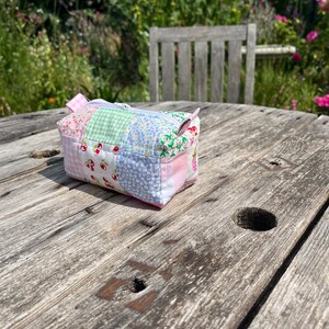 K&ouml;nnte beinhalten: Eine kleine Patchwork-Stofftasche mit Rei&szlig;verschluss. Die Tasche besteht aus verschiedenen rosa, gr&uuml;nen und blauen Stoffen mit einem Blumenmuster. Die Tasche liegt auf einem Holztisch.