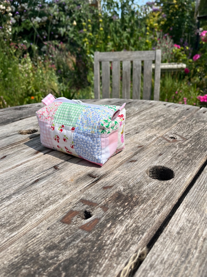 K&ouml;nnte beinhalten: Eine kleine Patchwork-Stofftasche mit Rei&szlig;verschluss. Die Tasche besteht aus verschiedenen rosa, gr&uuml;nen und blauen Stoffen mit einem Blumenmuster. Die Tasche liegt auf einem Holztisch.