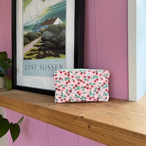 May include: A pink cosmetic bag with a pattern of red strawberries and green leaves. The bag is rectangular and sits on a wooden shelf. A framed poster of East Sussex is in the background.
