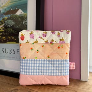 May include: A patchwork quilted pouch. The top section has a white background with pink strawberries. The middle section is peach with small orange fruit. The bottom section is blue gingham, and the base is pink.