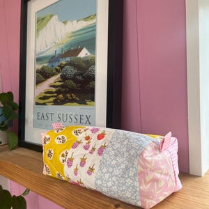 May include: A rectangular, quilted pencil case with a patchwork design. The case features sections of yellow with a floral pattern, strawberries, blue floral fabric, and pink floral fabric. A framed print of East Sussex is in the background.