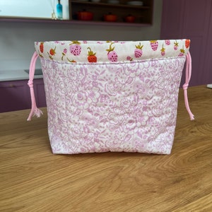 Könnte beinhalten: Ein gesteppter Kordelzugbeutel mit einem Blumenmuster in Weiß und Hellviolett. Der obere Rand hat einen Erdbeerdruck. Rosa Kordelzüge sind sichtbar. Die Tasche steht auf einer Holzoberfläche.