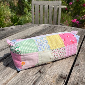 May include: A patchwork fabric pencil case with a zipper closure. The case is made of different coloured fabrics with floral and geometric patterns. The case is sitting on a wooden table.