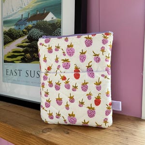 May include: A quilted white pouch featuring a strawberry pattern in pink and red, with green leaves. It has a purple zip and a small purple tab. The background includes a framed print and a pink wall.