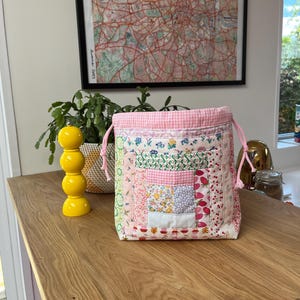 May include: A pink and white patchwork drawstring bag with a gingham top. The bag is made of various fabrics with floral and geometric patterns.