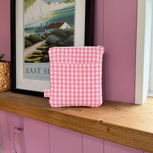 May include: A pink and white gingham pattern quilted pouch with a zip closure. The rectangular pouch has a small pink tag. The background features a framed landscape print and a wooden shelf. The print reads "East Sussex".