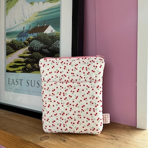 Può includere: Un astuccio trapuntato bianco con un motivo di ciliegie rosse. Ha una cerniera rosa e una piccola tasca frontale. Un piccolo cartellino rosa e bianco a scacchi è attaccato al lato.