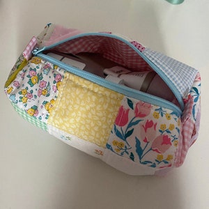 K&ouml;nnte beinhalten: Eine Rei&szlig;verschlusstasche aus Patchwork-Stoff mit einem hellblauen Rei&szlig;verschluss und einem Blumendruck. Die Tasche ist offen und enth&auml;lt eine wei&szlig;e und rosa Tube Lotion.