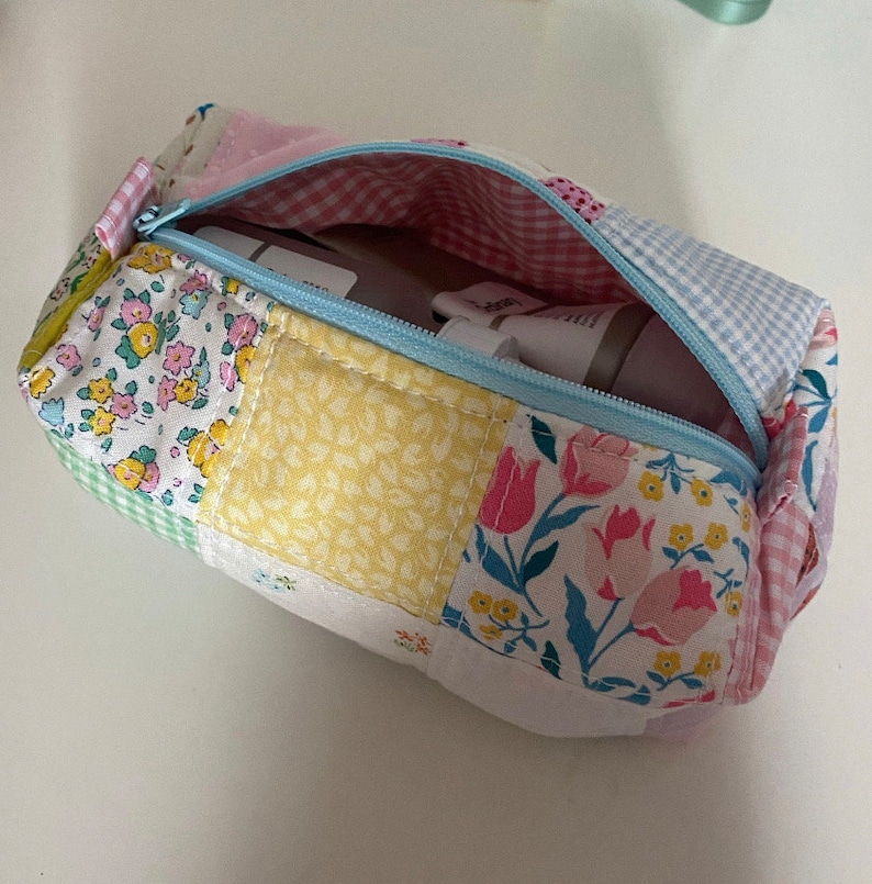 K&ouml;nnte beinhalten: Eine Rei&szlig;verschlusstasche aus Patchwork-Stoff mit einem hellblauen Rei&szlig;verschluss und einem Blumendruck. Die Tasche ist offen und enth&auml;lt eine wei&szlig;e und rosa Tube Lotion.