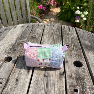 K&ouml;nnte beinhalten: Eine kleine, gepatchte Stofftasche mit Rei&szlig;verschluss in einem rosa, gr&uuml;nen und blauen Blumenmuster. Die Tasche liegt auf einem verwitterten Holztisch.
