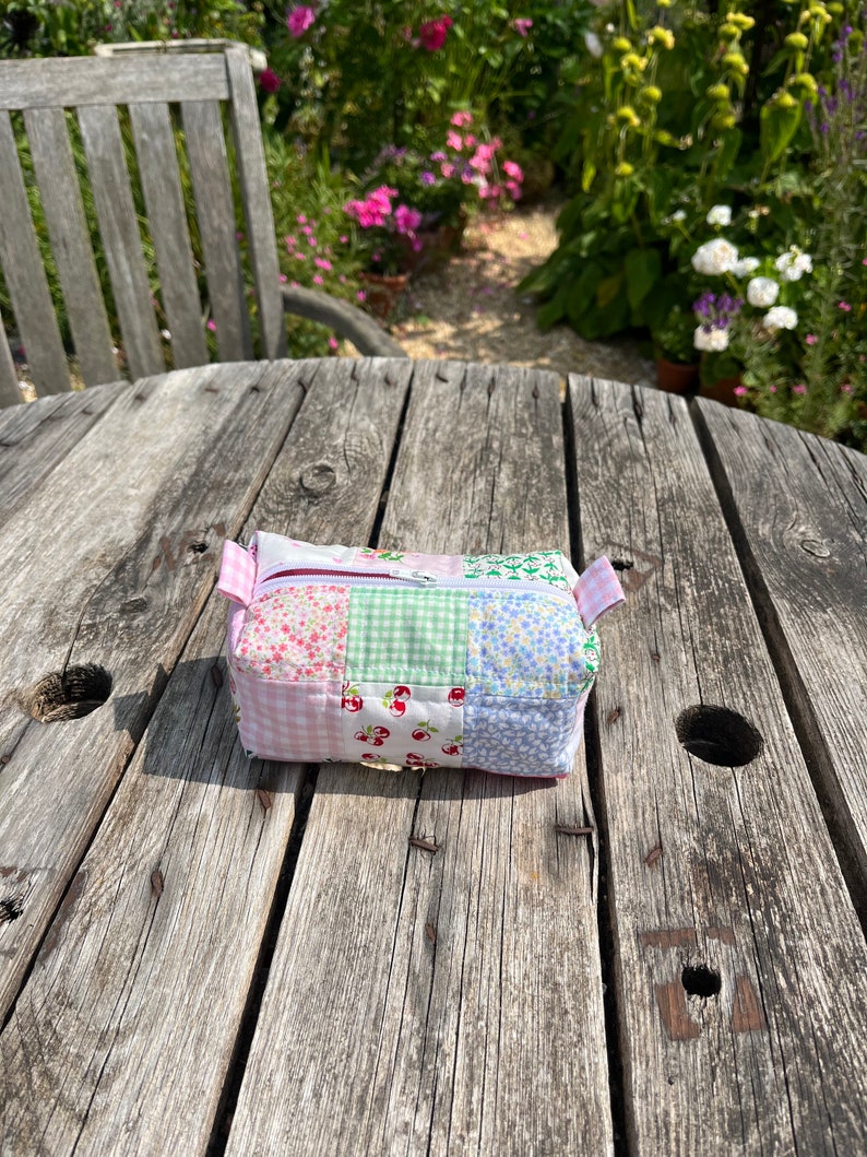 K&ouml;nnte beinhalten: Eine kleine, gepatchte Stofftasche mit Rei&szlig;verschluss in einem rosa, gr&uuml;nen und blauen Blumenmuster. Die Tasche liegt auf einem verwitterten Holztisch.