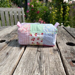 K&ouml;nnte beinhalten: Eine kleine, gepatchte Stofftasche mit Rei&szlig;verschluss und verschiedenen Blumen- und Karomustern in Rosa, Blau und Gr&uuml;n. Die Tasche liegt auf einer verwitterten Holzoberfl&auml;che.