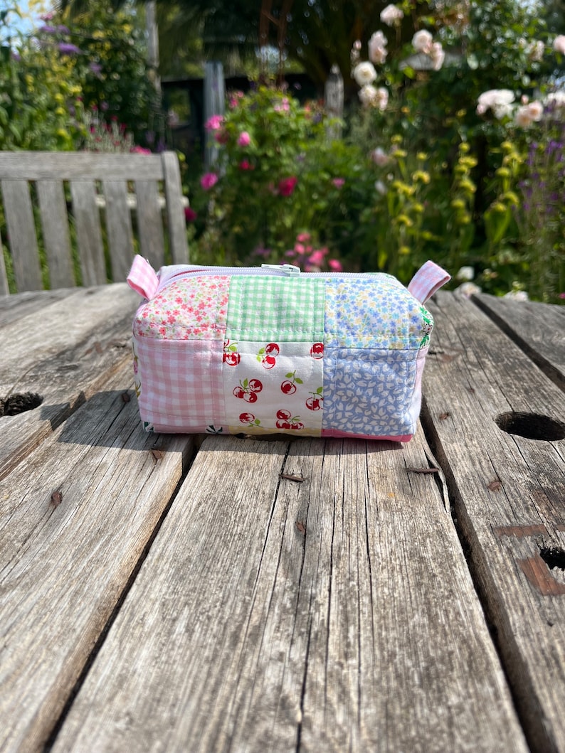 K&ouml;nnte beinhalten: Eine kleine, gepatchte Stofftasche mit Rei&szlig;verschluss und verschiedenen Blumen- und Karomustern in Rosa, Blau und Gr&uuml;n. Die Tasche liegt auf einer verwitterten Holzoberfl&auml;che.