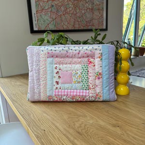 May include: A pink and blue patchwork quilt-style zippered pouch with a floral pattern. The pouch is made of various fabrics and has a square shape.