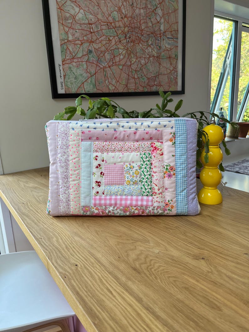 May include: A pink and blue patchwork quilt-style zippered pouch with a floral pattern. The pouch is made of various fabrics and has a square shape.