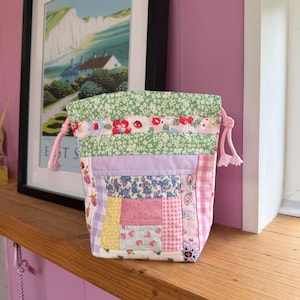 May include: A fabric drawstring pouch featuring a patchwork design with various floral, gingham, and strawberry patterns in shades of pink, green, yellow, and lavender. The bag has a green floral top border and light pink knotted drawstring cords.