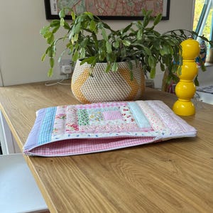 May include: A pink and blue patchwork fabric laptop sleeve with a zipper closure. The sleeve is lying on a wooden table.