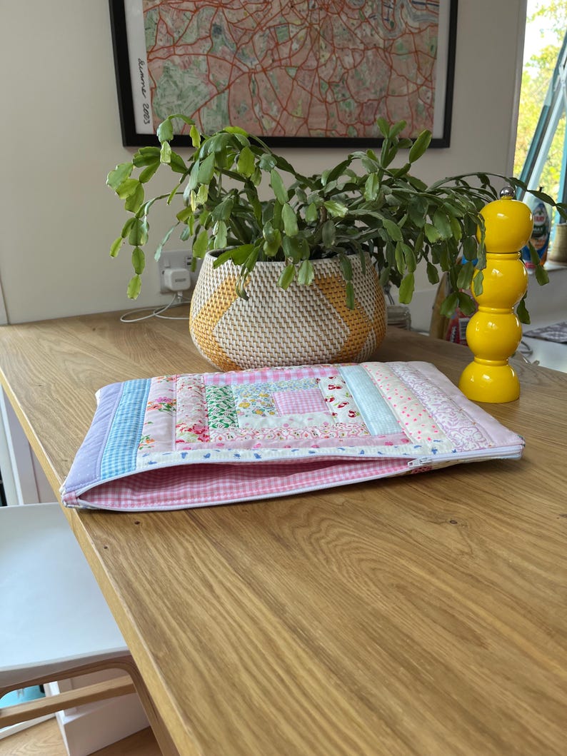 May include: A pink and blue patchwork fabric laptop sleeve with a zipper closure. The sleeve is lying on a wooden table.