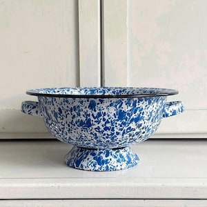 May include: A round enamel colander featuring a white base with a blue speckled pattern. The bowl has two side handles, a pedestal base, and a black rim. Numerous small circular drainage holes are distributed across the body of the bowl.