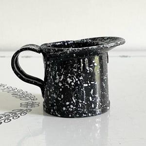 May include: A small, black and white speckled enamelware pitcher with a handle. The pitcher has a flared spout and a capacity marking on the side. The speckled pattern is white on a black background.