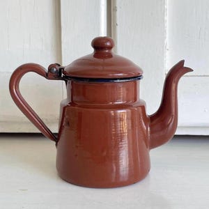 May include: A vintage, brown enamel teapot with a lid and curved handle. The teapot has a spout and a dark blue trim around the lid. The teapot is on a white surface.