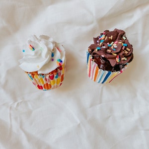 Puede incluir: Dos cupcakes sobre una superficie blanca. Uno tiene glaseado blanco con chispas de arcoíris, el otro tiene glaseado de chocolate con chispas de arcoíris. Ambos están en envoltorios de cupcakes a rayas de colores.