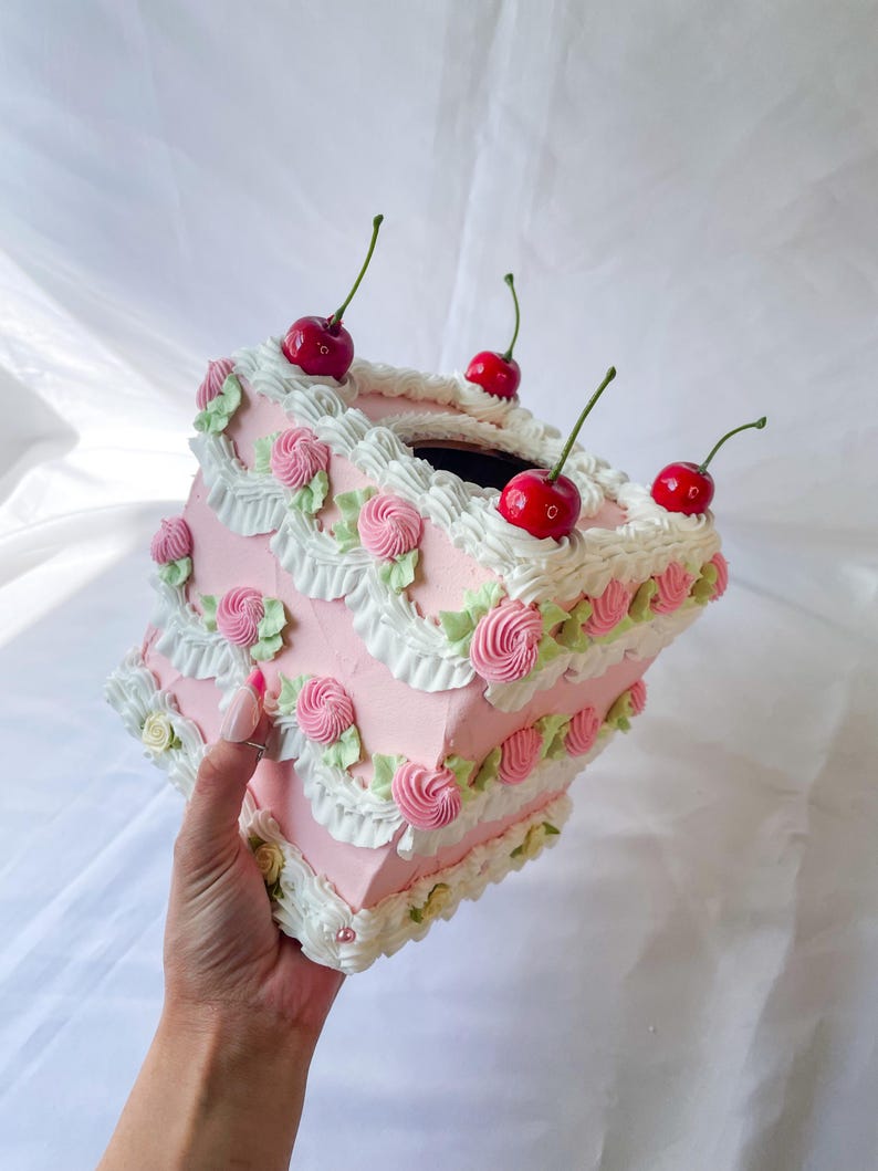 May include: A pink and white cake-shaped tissue box holder with white frosting and red cherries on top.