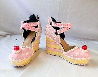 May include: A pair of whimsical wedge heels designed to resemble slices of cake. The shoes feature a yellow base, pink frosting, white floral accents, and a red cherry on top. The heels have a black strap with a pink bow.